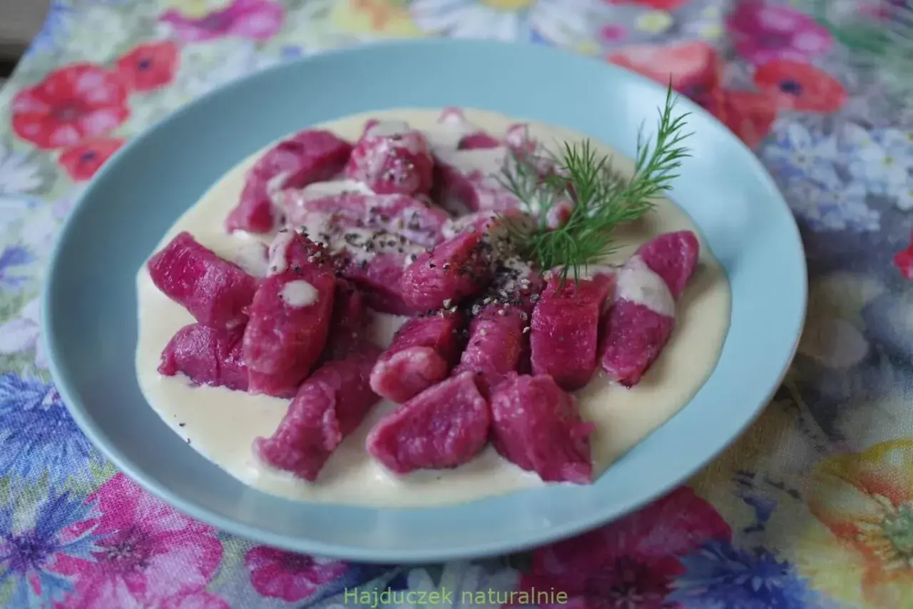 Różowe gnocchi buraczane w kremowym sosie, posypane pieprzem i udekorowane gałązką koperku, na talerzu z kwiecistym obrusem.