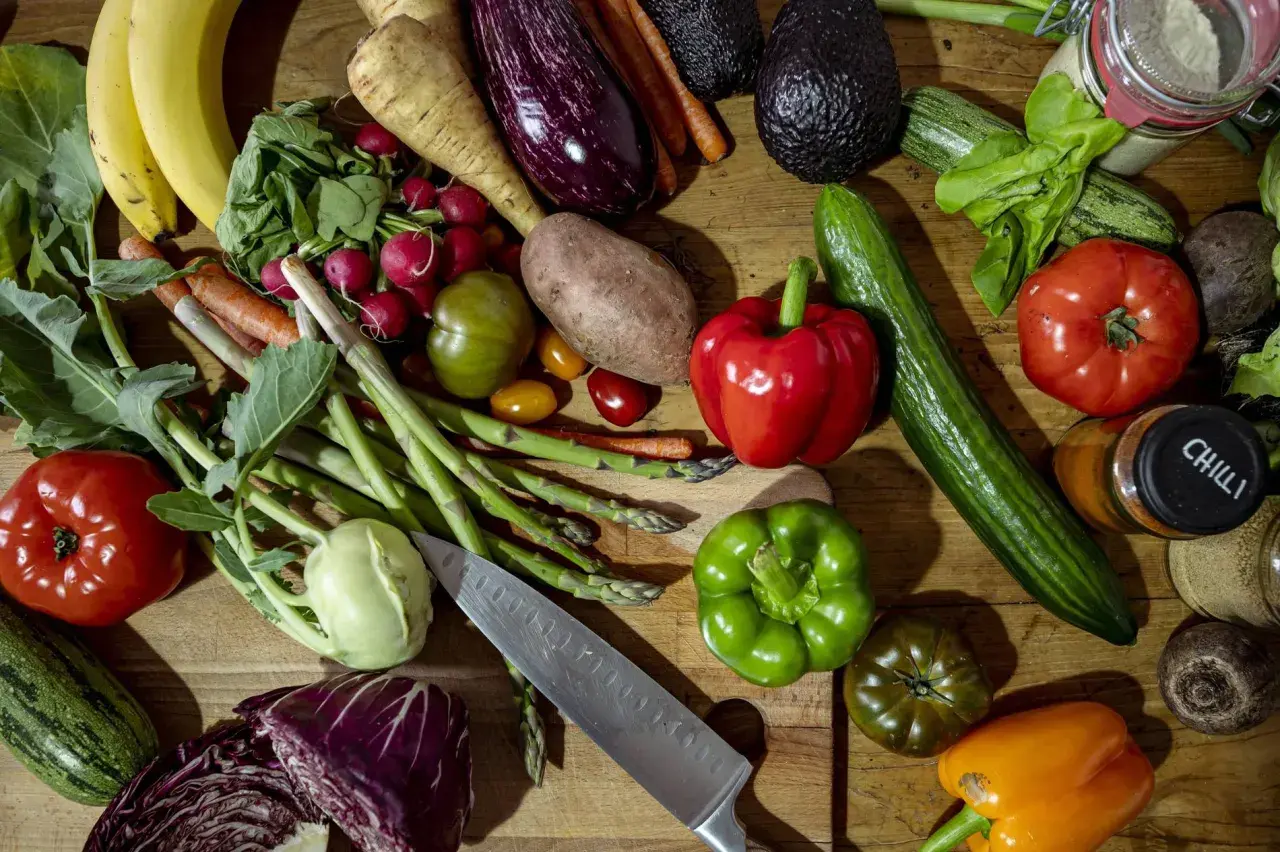 Buntes Arrangement von frischem Gemüse und Obst, darunter Bananen, Paprika, Tomaten, Gurken, Auberginen und Spargel. Ein Messer liegt daneben. Alles für gesundes Essen.