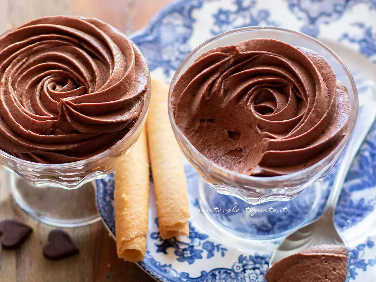Due coppette di mousse al cioccolato ricetta classica, guarnite con rotoli di wafer e cuoricini di cioccolato.