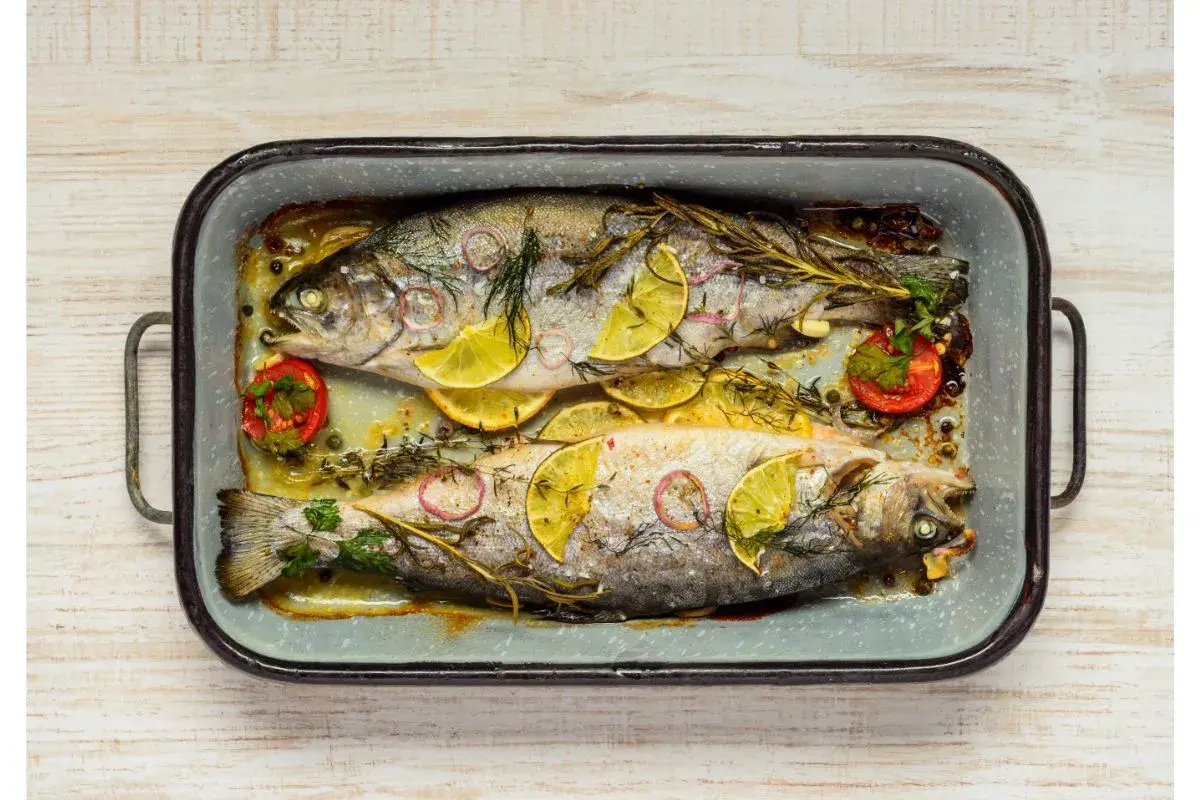 Deux truites au four, garnies de rondelles de citron, d'oignon rouge, de tomates cerises et d'herbes aromatiques, dans un plat en émail.