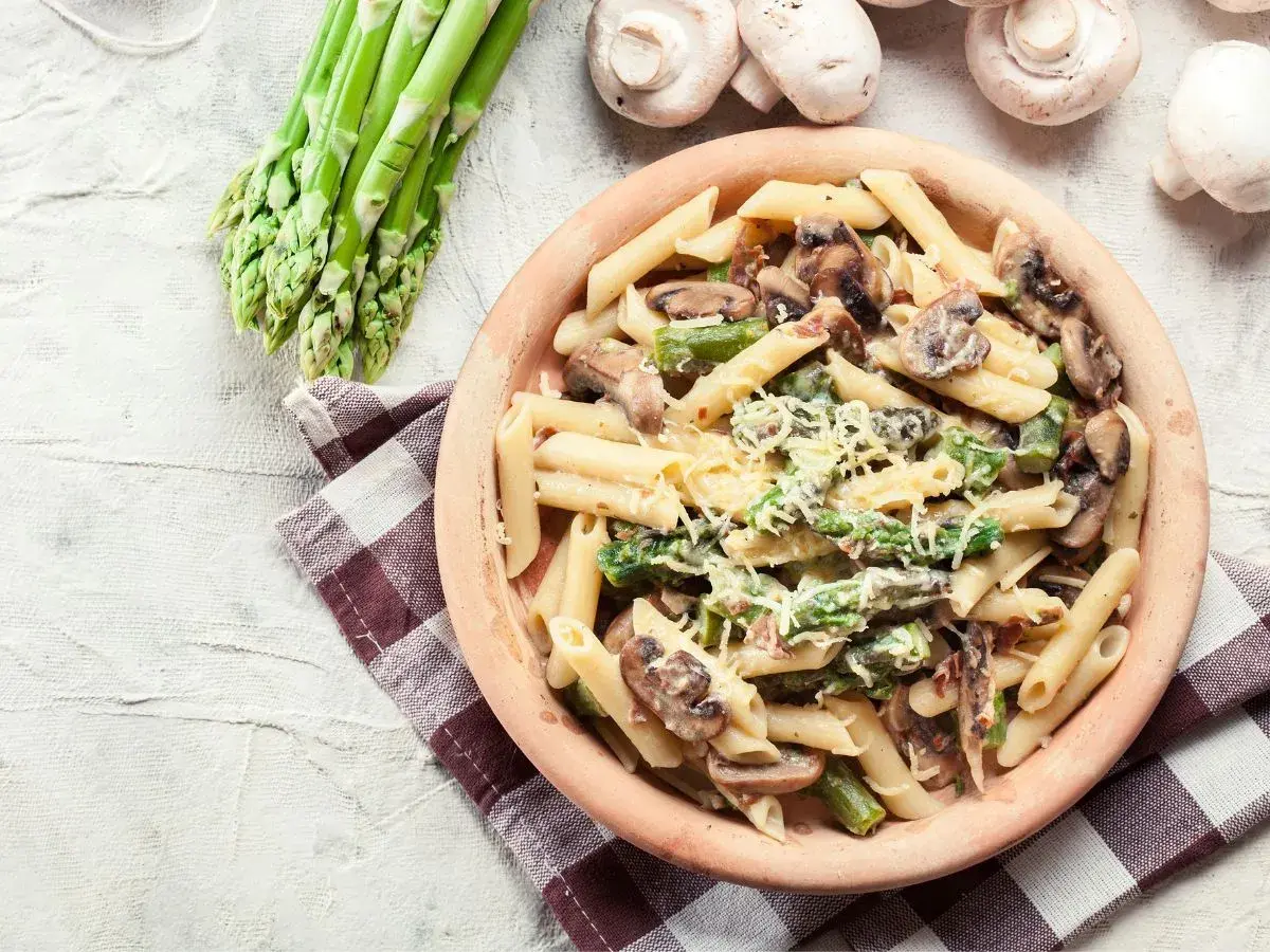 Penne z kremowym sosem, szparagami i pieczarkami, posypane parmezanem. Obok pęczek świeżych szparagów i kilka pieczarek.