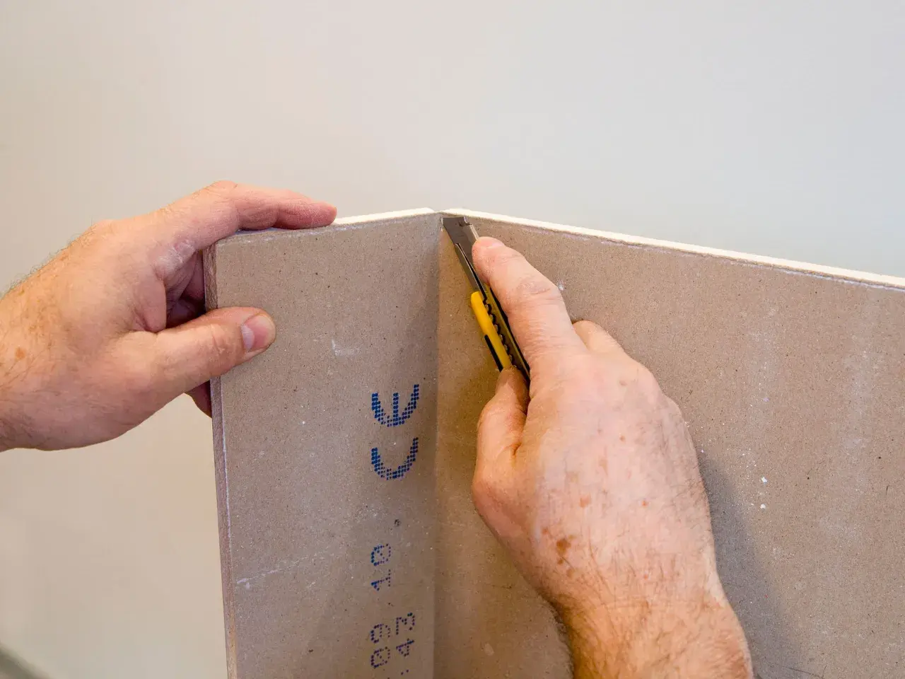 Mani che tengono un cartongesso mentre un taglierino viene usato per tagliare il materiale. Impara come tagliare il cartongesso con precisione.