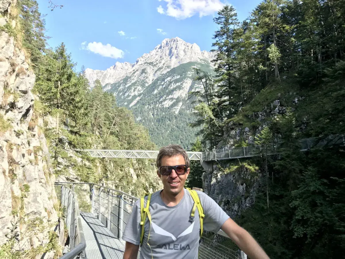 Mann auf einem Steg in der Leutascher Geisterklamm, umgeben von Felsen und Bäumen, mit majestätischen Bergen im Hintergrund.