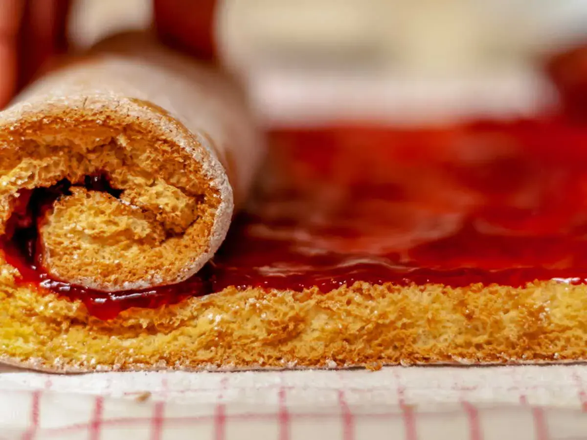Un rouleau de génoise moelleuse, saupoudré de sucre glace, est en train de rouler sur une couche de confiture de fraises éclatante.