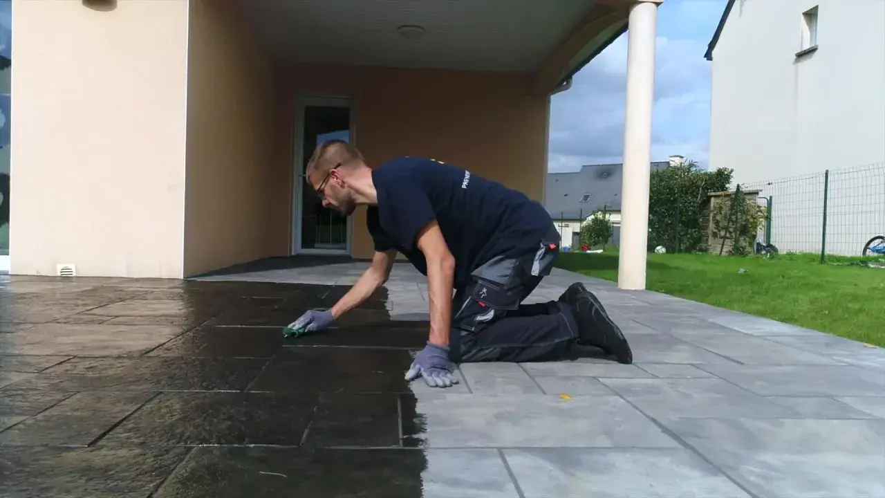 Un homme agenouillé effectue le nettoyage de pierre naturelle extérieur avec une éponge, redonnant son éclat à la terrasse.