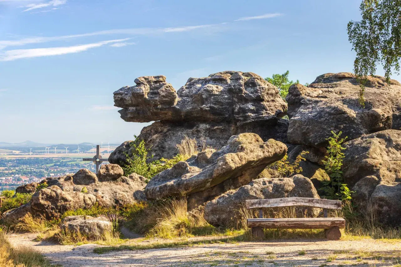 Ein Holzbank lädt zum Verweilen ein, mit Blick auf bizarre Felsen im Zittauer Gebirge und eine Stadt in der Ferne.