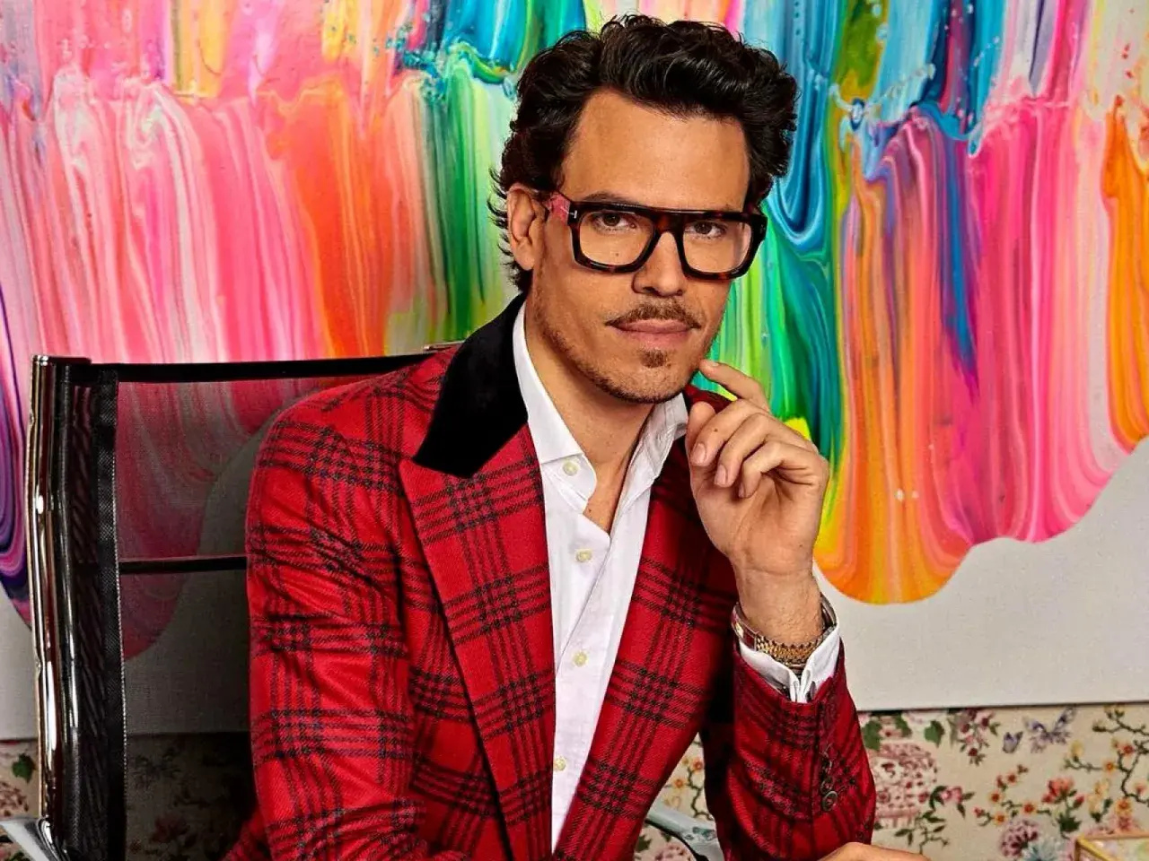 Juan Avellaneda, con su característico traje a cuadros rojos y gafas llamativas, posa con una mano en la barbilla frente a un vibrante cuadro abstracto.
