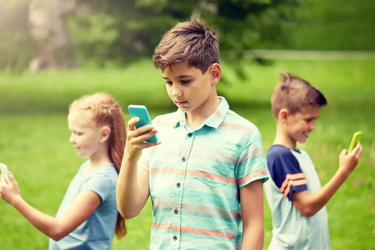 Dzieci w parku korzystają z telefonów. Chłopiec w centrum z niebieskim telefonem, dziewczynka po lewej z białym, a chłopiec po prawej z zielonym. Bezpieczne surfowanie dzięki Google VPN.