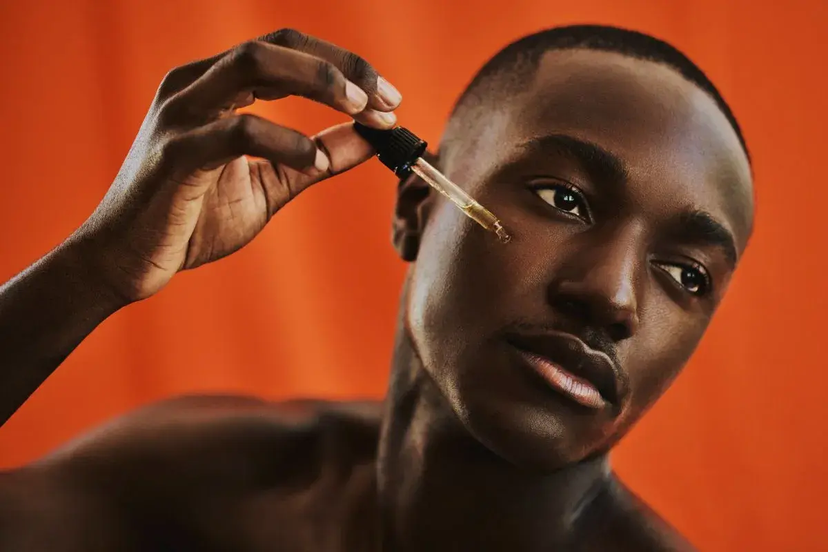 Uomo con pelle scura applica un siero con contagocce, parte della sua beauty routine viso.