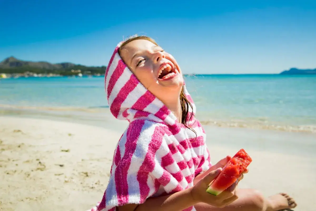 Niña riendo en la playa con una toalla a rayas y sandía, perfecta comida para llevar a la playa con niños.