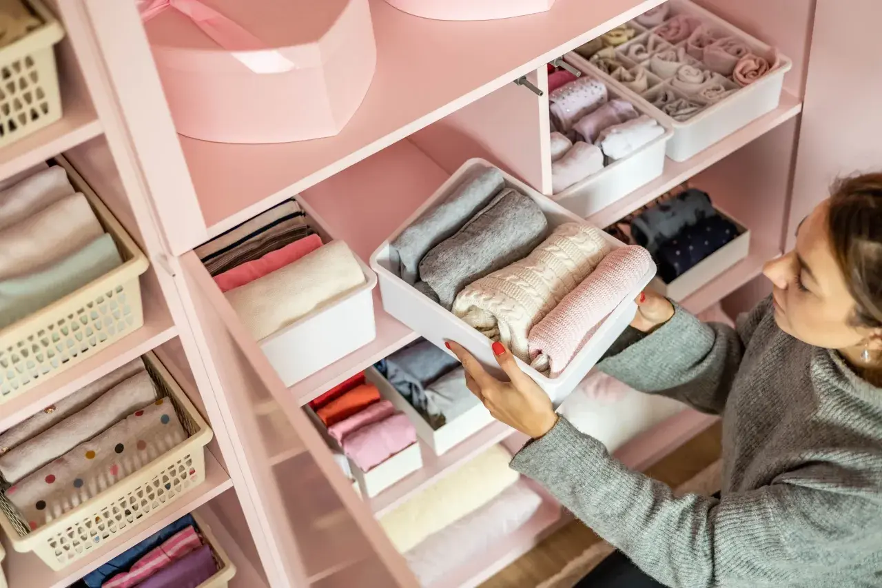 Frau sortiert Kleidung in einem rosa Kleiderschrank. So organisiere ich meinen Kleiderschrank: gefaltete Pullover und Socken in Behältern.