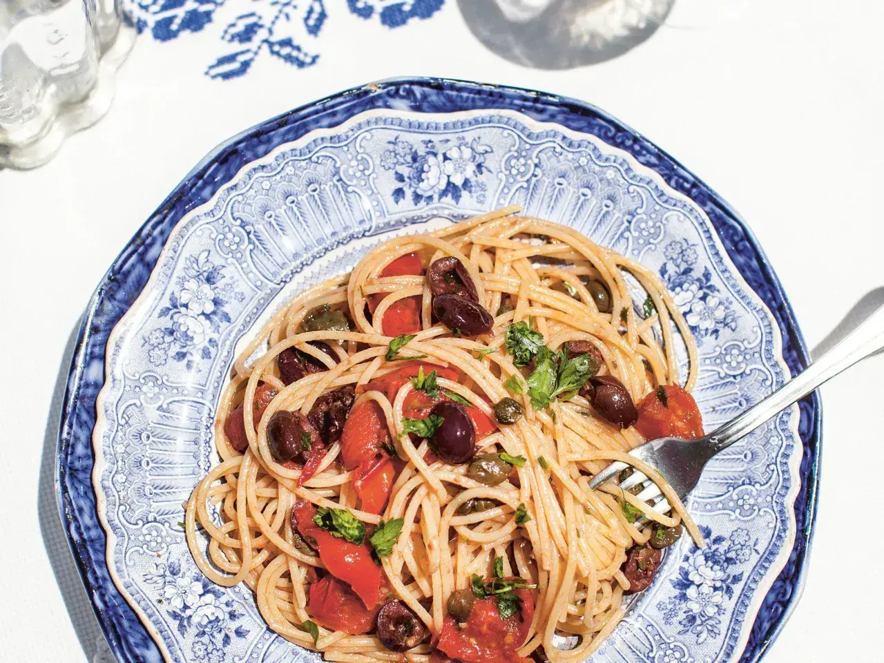 Spaghetti Puttanesca mit Oliven, Kapern und Tomaten auf einem blau-weißen Teller.