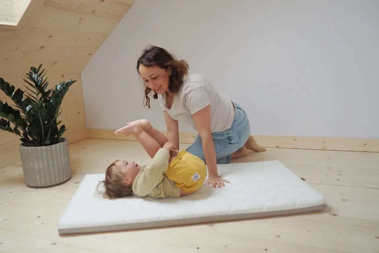 Mama bawi się z dzieckiem na miękkim materacyku do łóżeczka. Radosny maluch leży na plecach, a mama klęczy obok, uśmiechając się.