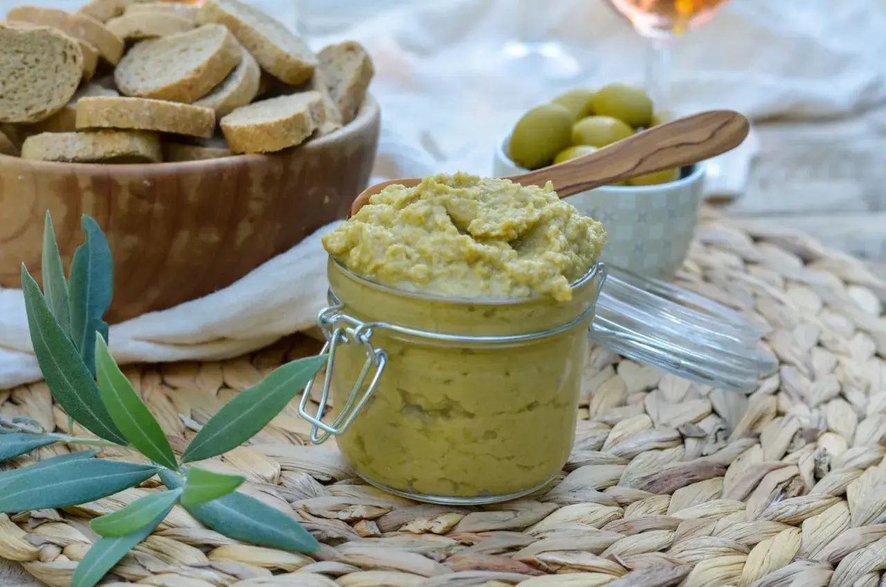 Un pot de tapenade verte, garni d'une cuillère en bois, est prêt à être dégusté avec des toasts et des olives.