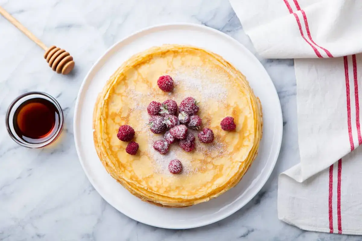 Ein Stapel goldbrauner Ofenpfannkuchen, garniert mit Himbeeren und Puderzucker. Daneben eine Schale Honig und ein Honiglöffel. Perfekt für den Thermomix!