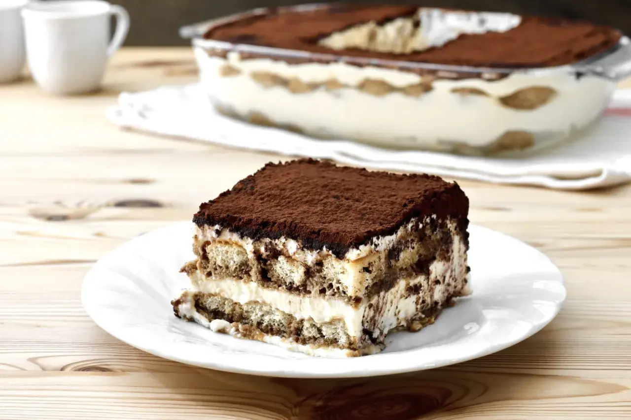 Una fetta di tiramisù su un piatto bianco, con la teglia sullo sfondo. La crema tiramisù perfetta, soffice e stratificata, è cosparsa di cacao.