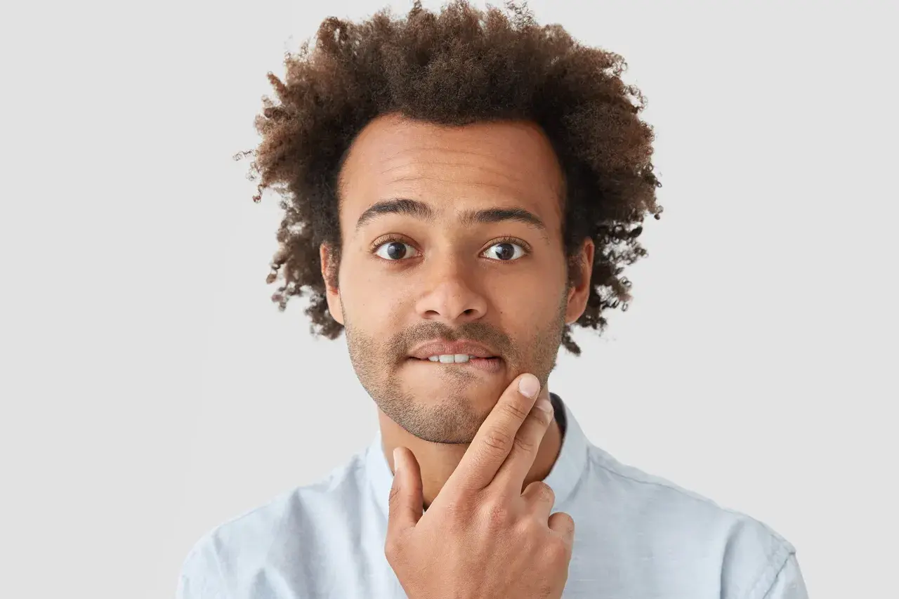 Uomo con capelli ricci e barba corta, con un dito sul mento, espressione pensierosa, come se avesse un sapore acido in bocca.