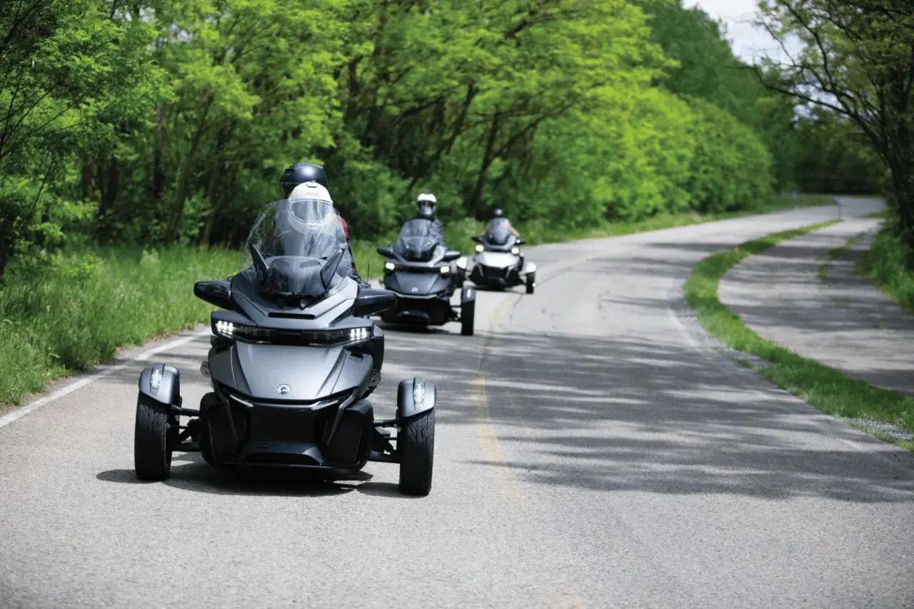 Trzy pojazdy typu Can-Am Spyder jadące po krętej drodze wśród zieleni. Ich nowoczesny design i dynamiczna jazda tworzą wrażenie efektu shimmy.