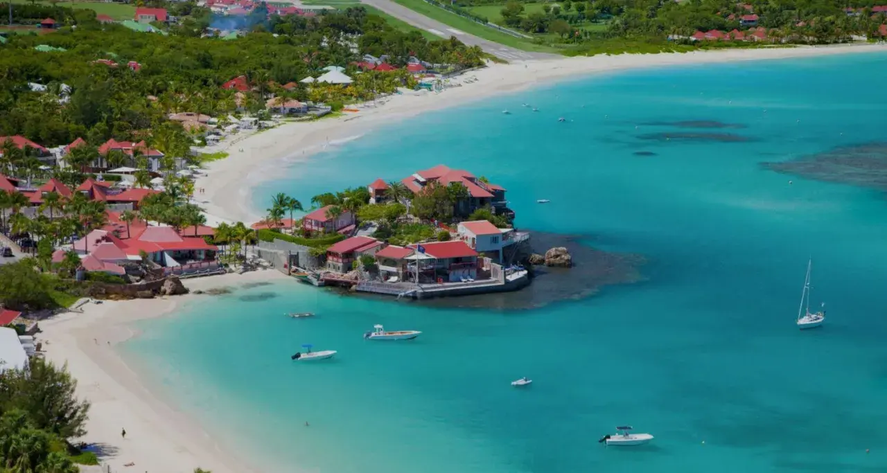 Vue aérienne de Saint-Barthélemy, avec ses plages de sable blanc, ses eaux turquoise et ses villas aux toits rouges. Un voilier navigue paisiblement.