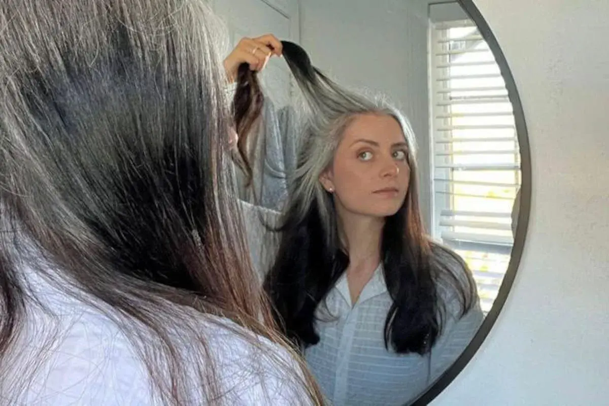 Mujer se mira al espejo, su cabello es una mezcla de negro y canas amarillas, un estilo natural y único.