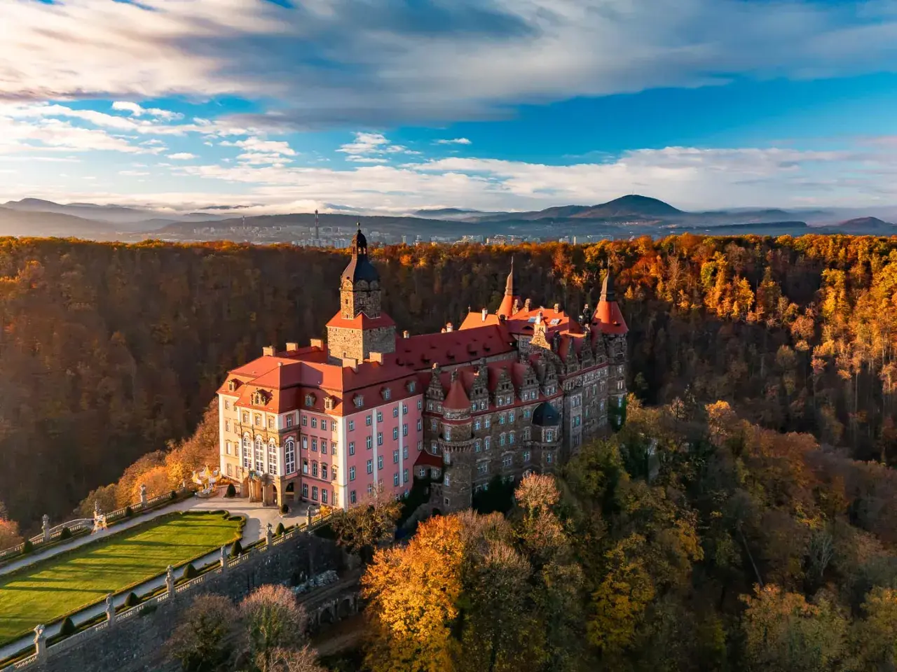 Malowniczy hotel zamek otoczony jesiennym lasem, z wieżą i czerwonymi dachami, na tle gór i błękitnego nieba.