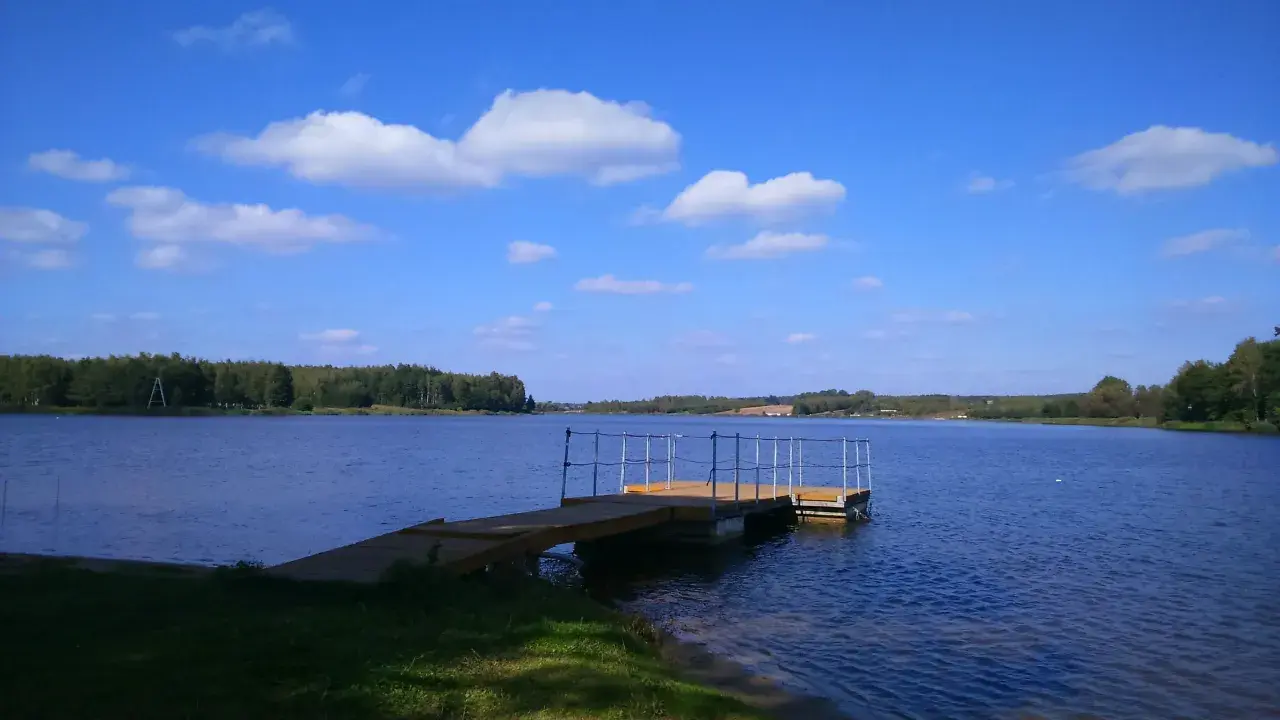 Drewniana przystań na spokojnym zalewie Drzewica. Błękitne niebo z białymi chmurami nad taflą wody i zielonymi lasami w tle.