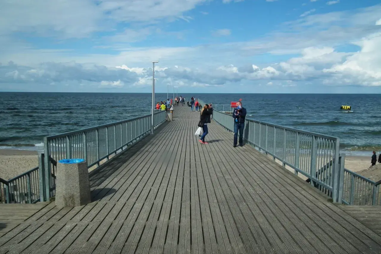 Ludzie spacerują po drewnianym deptaku nad morzem. Słońce przebija się przez chmury, a fale delikatnie uderzają o brzeg.