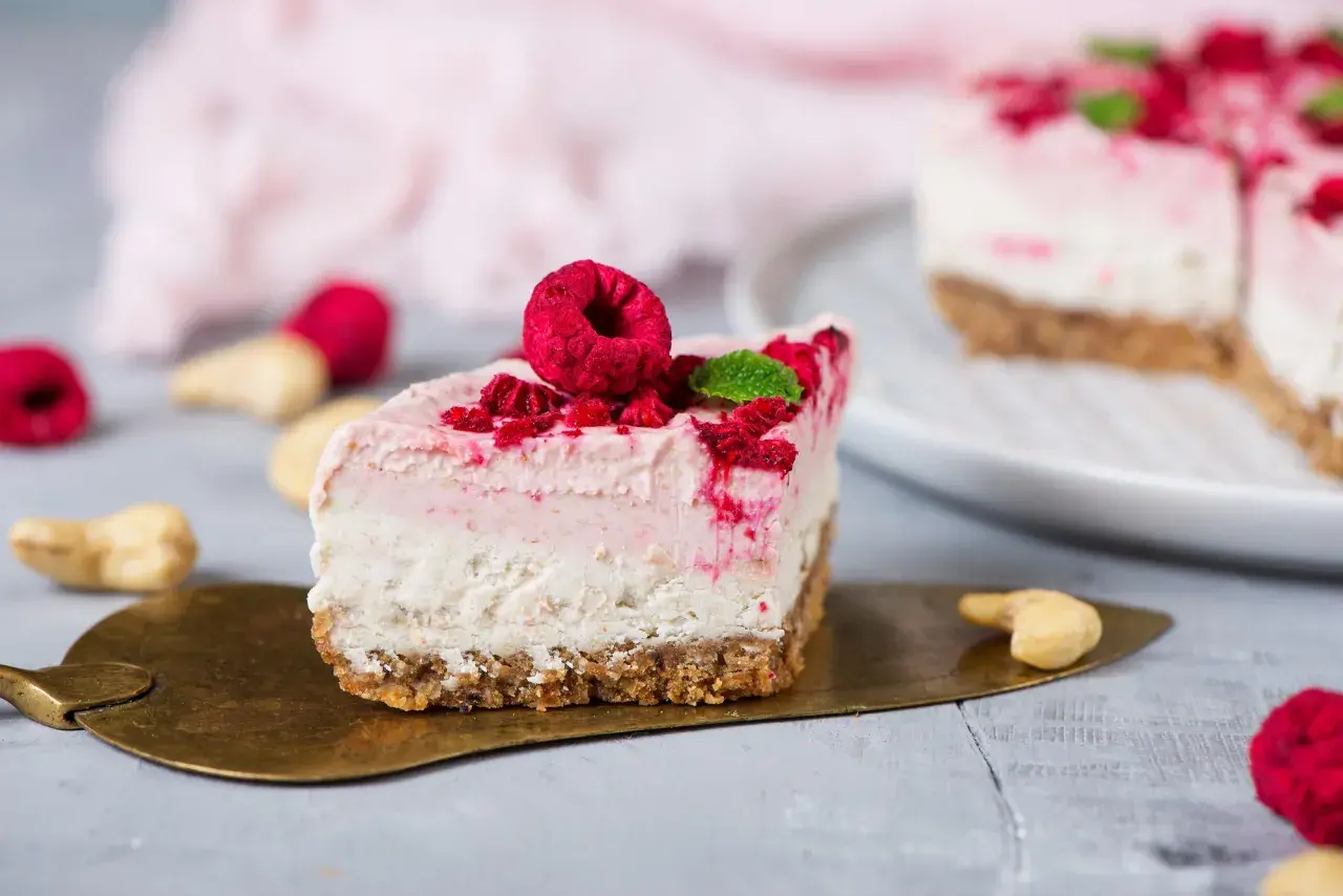 Ein Stück Himbeer Cheesecake mit frischen Himbeeren und Minze auf einem goldenen Tortenheber.