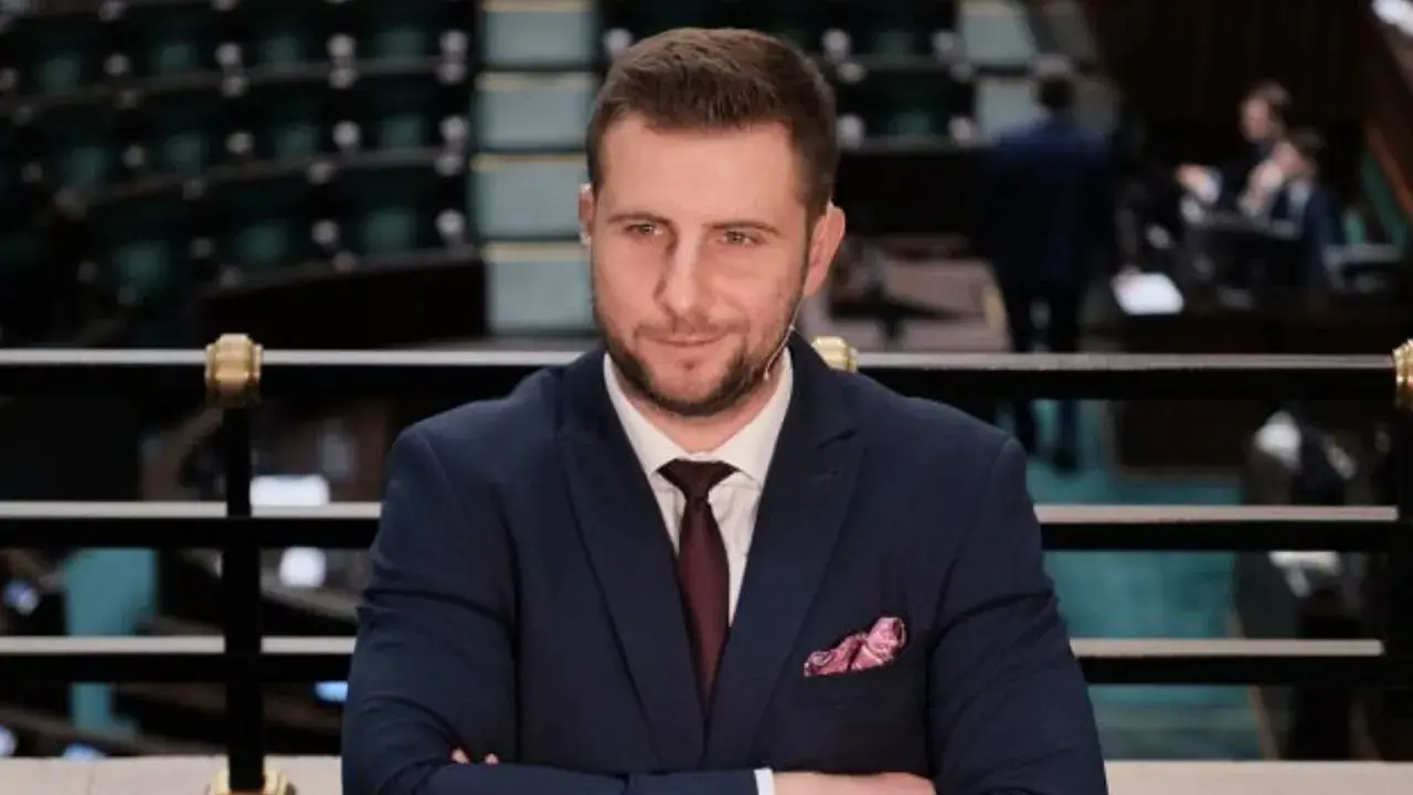 Mężczyzna w garniturze z krawatem i poszetką, siedzi na tle sali sejmowej.