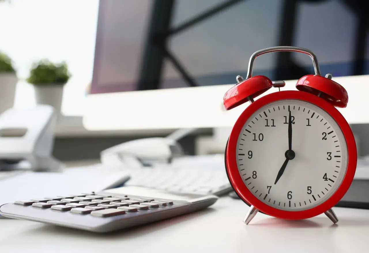 Sulla scrivania, un orologio rosso segna le 7. La calcolatrice e il computer suggeriscono che il giorno di riposo viene pagato, ma il lavoro attende.