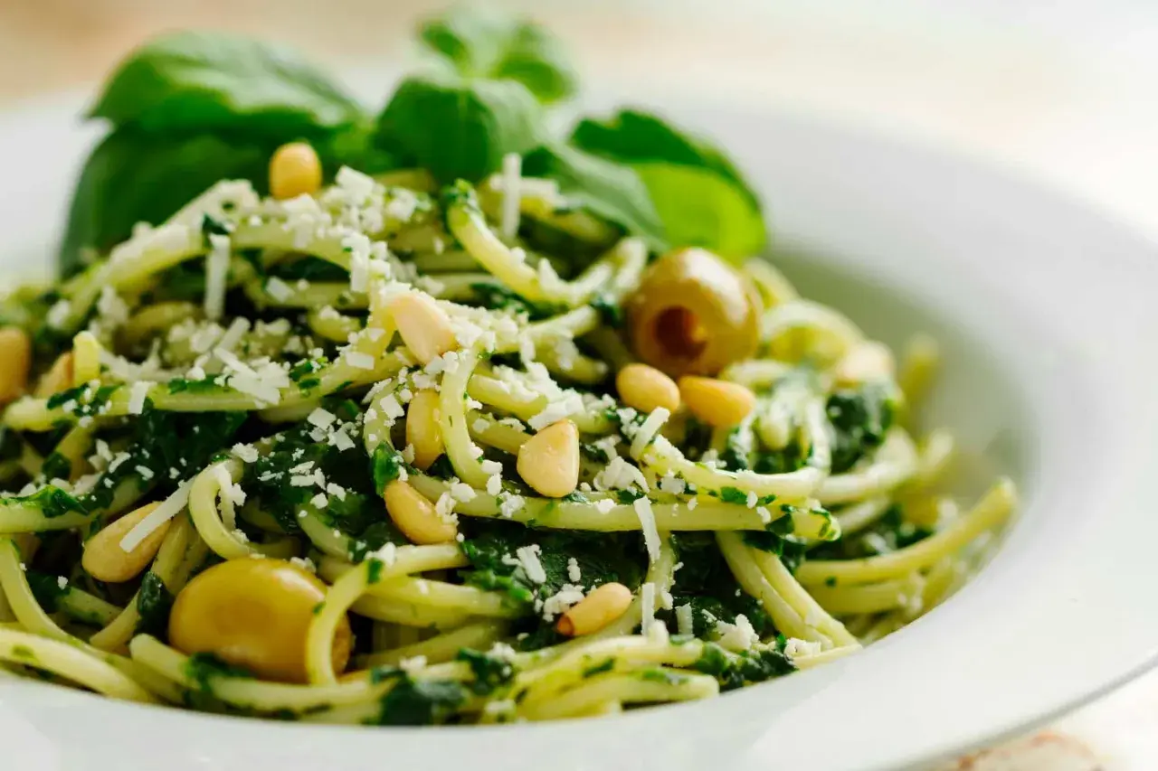 Pasta con pesto de pistacho, piñones, aceitunas verdes y queso rallado, decorada con hojas de albahaca fresca.