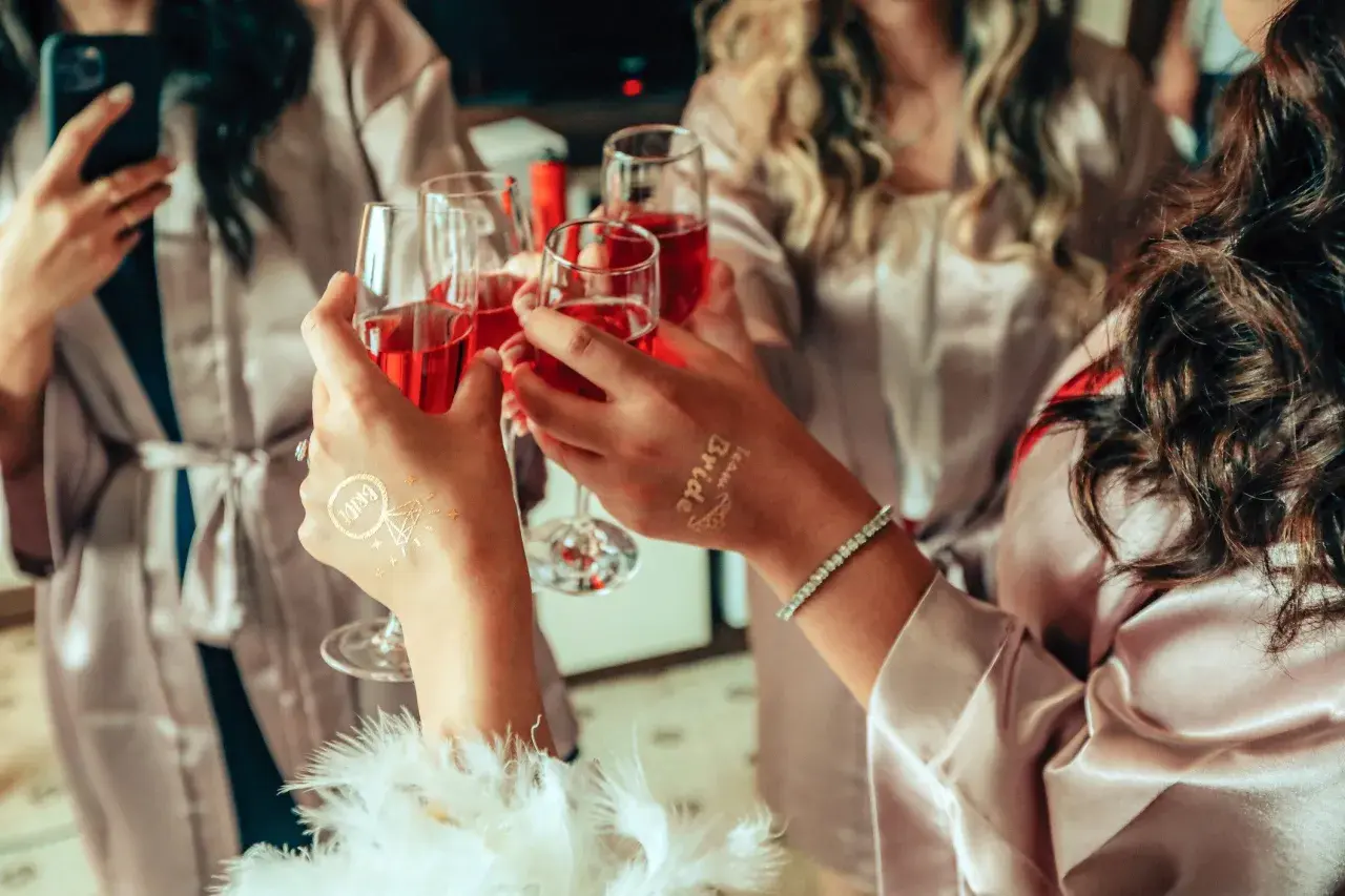 Dłonie w szlafroczkach unoszą kieliszki z czerwonym napojem, na jednej widoczny tatuaż "Bride".