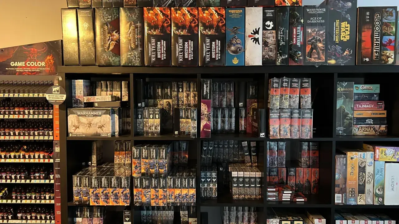 Półki pełne pudełek z grami i figurkami, w tym wiele z serii Warhammer. Widać tu zapowiedź przygód Gotreka i Felixa, a także inne tytuły.