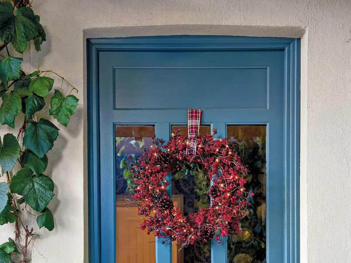 Corona navideña roja con luces y piñas colgada en una puerta azul.
