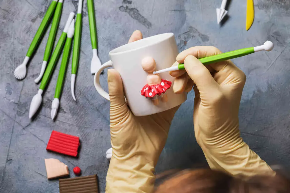 Mani guantate creano una bambolina con pasta fimo su una tazza. Tanti strumenti e colori per idee creative.