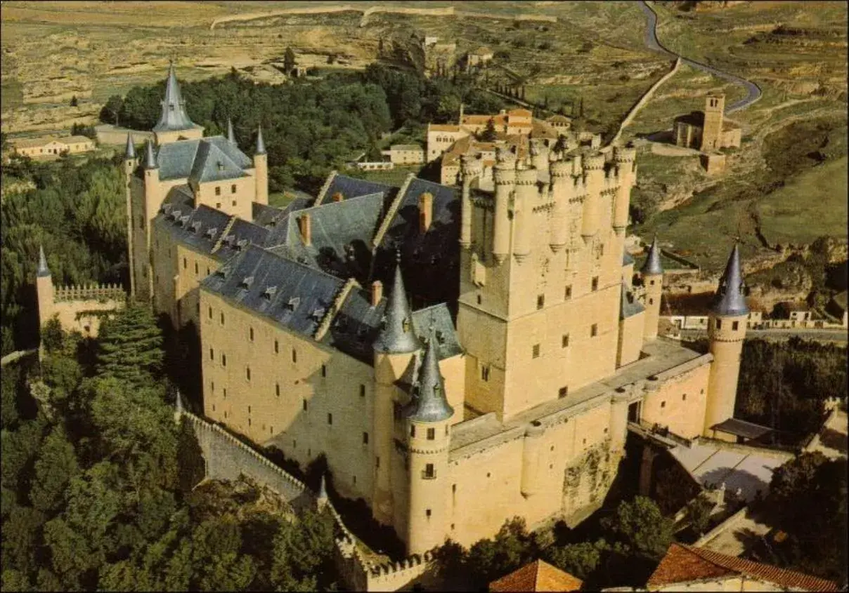Vista aérea del Alcázar de Segovia, con sus torres y tejados, rodeado de vegetación. La sala de los reyes se intuye en su imponente estructura.