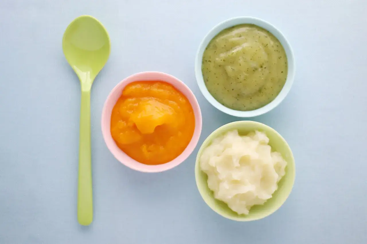 Drei Schälchen mit Brei (orange, grün, weiß) und ein Löffel. Ideal für die Beikosteinführung: ab wann Obstbrei Baby?