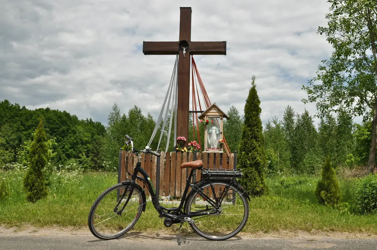 Rower przy kapliczce w Hajnówce, Białowieża. Drewniany krzyż z figurką Chrystusa i Matki Boskiej, otoczony zielenią i polnymi kwiatami.