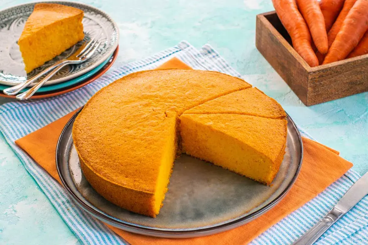 Una deliziosa torta alle carote, soffice e dorata, tagliata a fette su un piatto. Accanto, carote fresche in un cesto di legno.