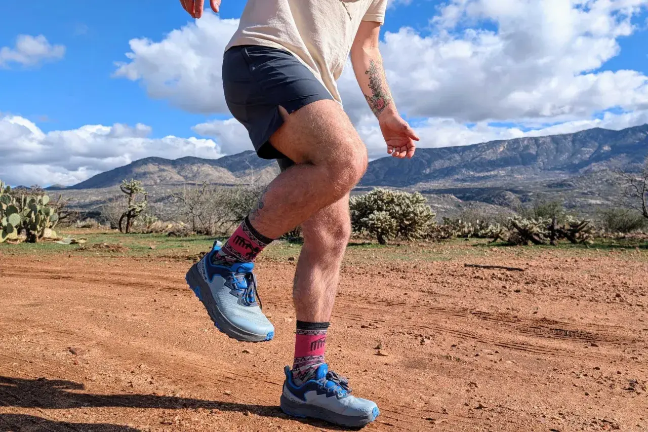 Runner's legs in Altra Timp 6 shoes and patterned socks, mid-stride on a dusty desert trail with mountains in the background.