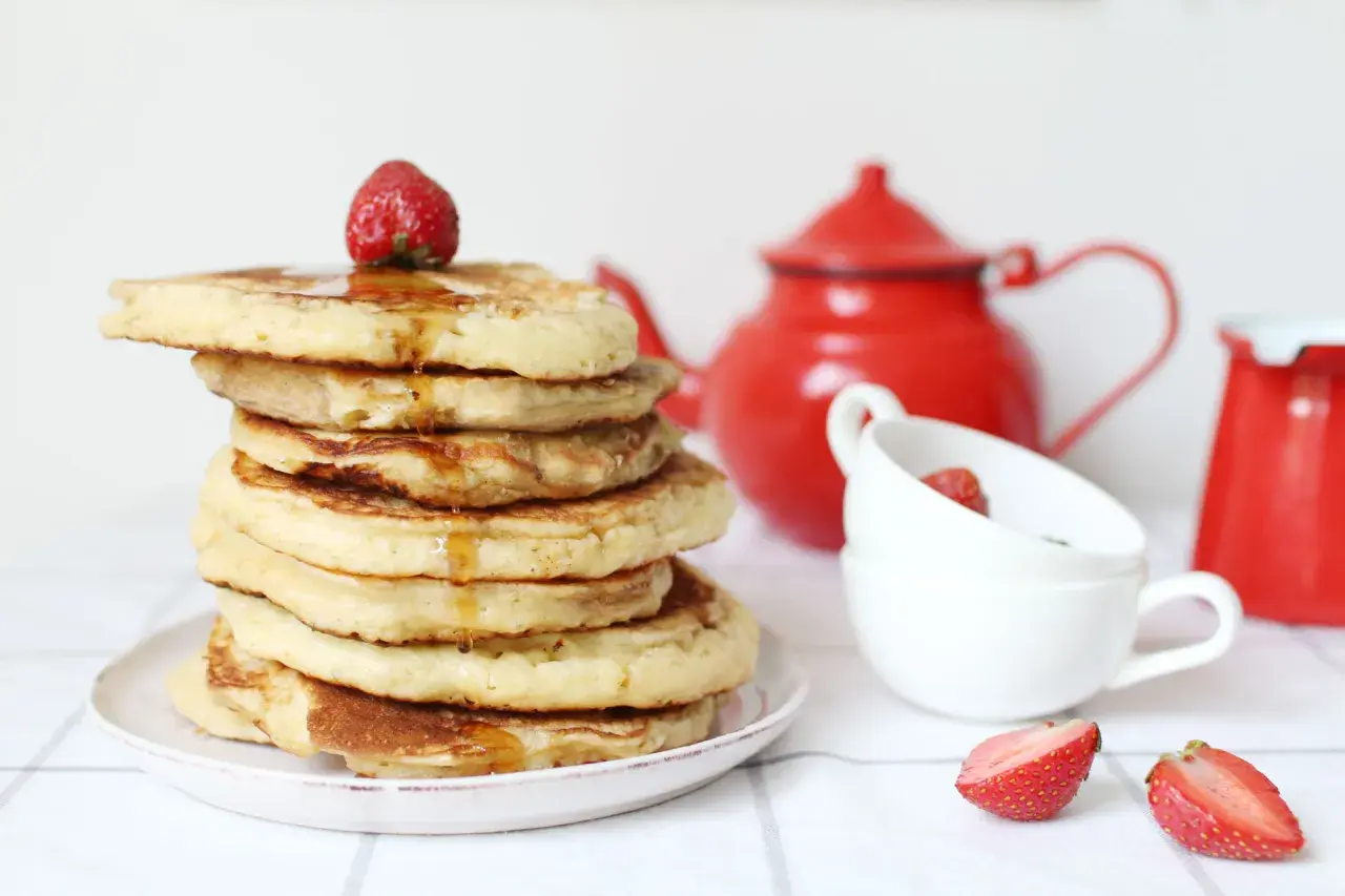 Pile de pancake sans gluten, nappés de sirop et garnis d'une fraise. Une théière rouge et des tasses complètent la scène.