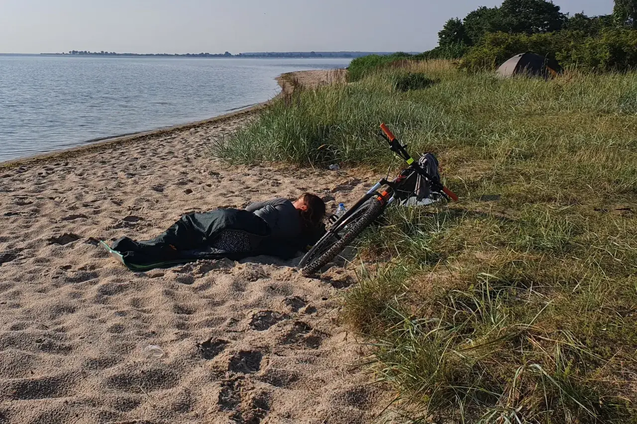 Osoba śpi na plaży obok roweru. W oddali widać wodę i drzewa. To dowód, że można spać na plaży.