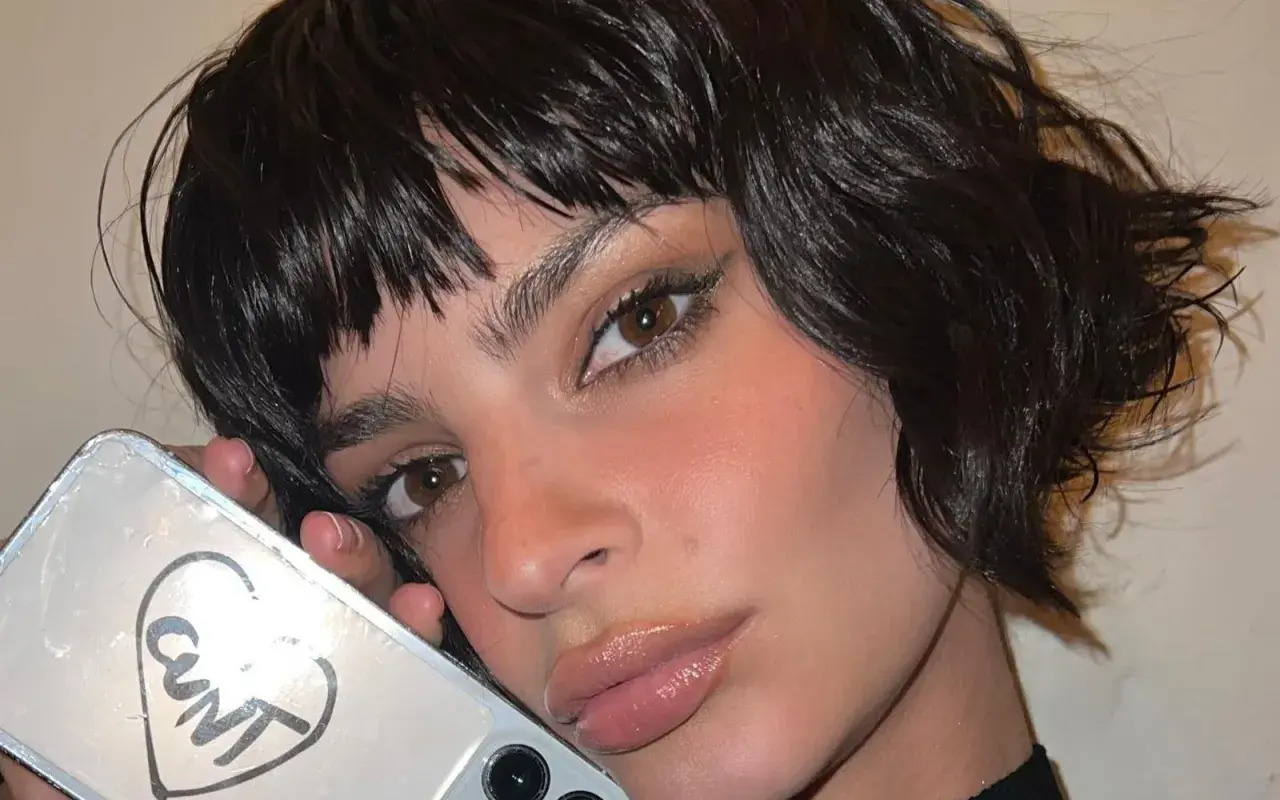 Primo piano di una donna con capelli scuri e umidi che le incorniciano il viso. Tiene in mano un telefono con un disegno a cuore e la scritta "CUNT".