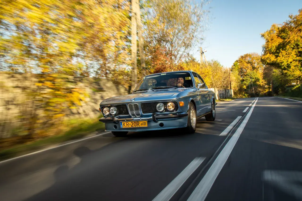 Błękitne BMW E9 z kierowcą jedzie jesienną drogą wśród kolorowych drzew.