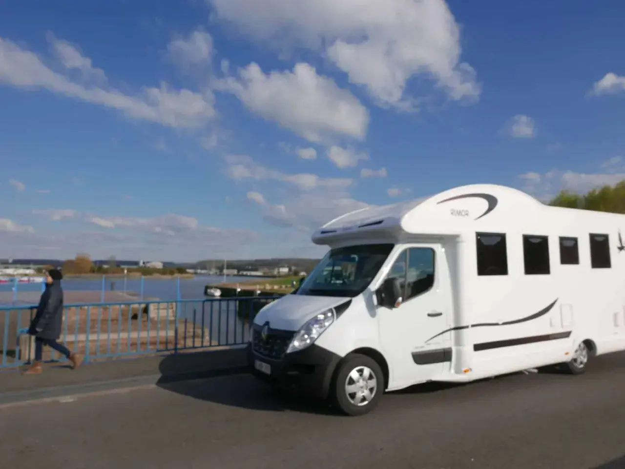 Un camping-car blanc roule sur un pont, avec une personne marchant à côté. Le ciel est bleu avec des nuages. Idéal pour la consommation camping-car.