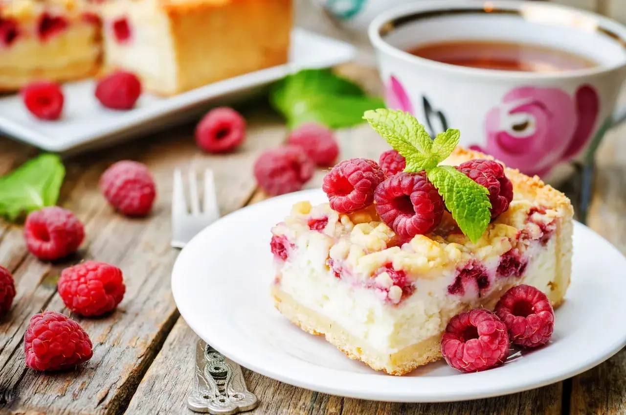 Malinowa chmurka z serem i malinami, udekorowana listkiem mięty, na talerzyku obok filiżanki herbaty.