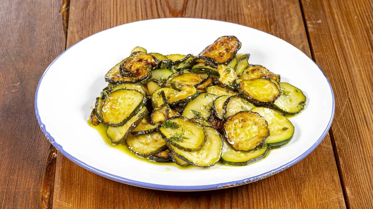 Piatto di zucchine alla poverella, fette sottili e dorate condite con olio e erbe aromatiche, su un piatto bianco con bordo blu.