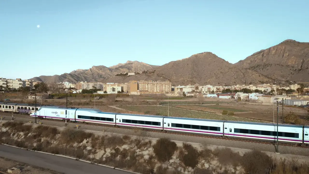 Tren AVE de Renfe, un moderno tren blanco con detalles morados, atraviesa un paisaje rural con montañas y edificios al fondo.