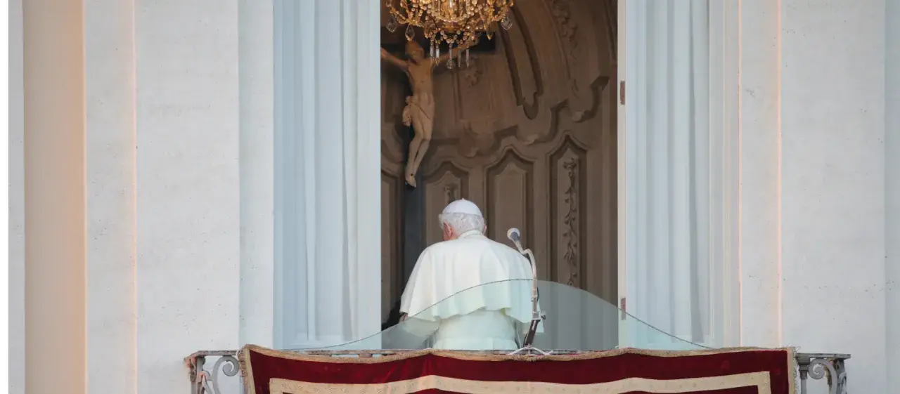 Der Papst steht auf dem Balkon, hinter ihm ein Kruzifix und ein Kronleuchter.
