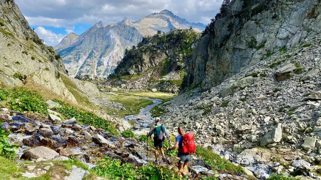 Hikers ascend a rocky path alongside a stream, with majestic mountains in the distance, on the Haute Route Pyrenees.
